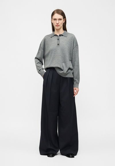 Γυναίκα με διαφανή γυαλιά, γκρι πουλόβερ πόλο με μακριά μανίκια, μαύρο πλισέ παντελόνι με φαρδιά ποδόγυρα και μαύρα παπούτσια, στεκόμενη με το ένα χέρι στην τσέπη.