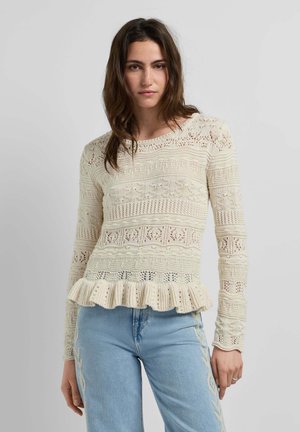 Femme portant un pull en maille crochet crème avec un ourlet à volants et un jean bleu clair avec des motifs brodés sur les côtés, debout devant un fond uni.