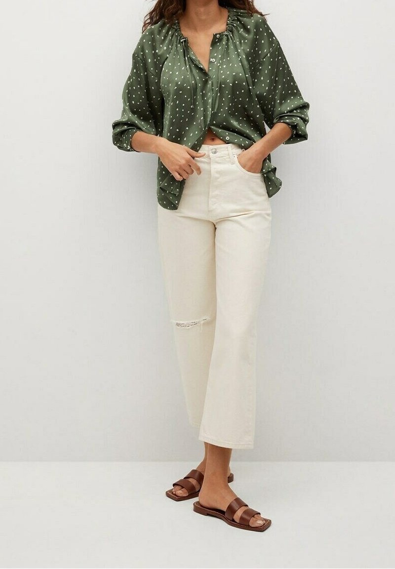 Blusa verde a pois, vestibilità ampia con scollatura arricciata, abbinata a jeans bianchi a vita alta, leggermente sfrangiati, e sandali scivolati marroni.