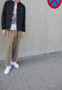 Černá lehká bunda, bílá květinová košile, béžové manšestrové kalhoty, bílé slip-on boty. Stojí opřený o betonovou zeď s dopravní značkou zákazu parkování.