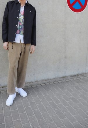 Černá lehká bunda, bílá květinová košile, béžové manšestrové kalhoty, bílé slip-on boty. Stojí opřený o betonovou zeď s dopravní značkou zákazu parkování.