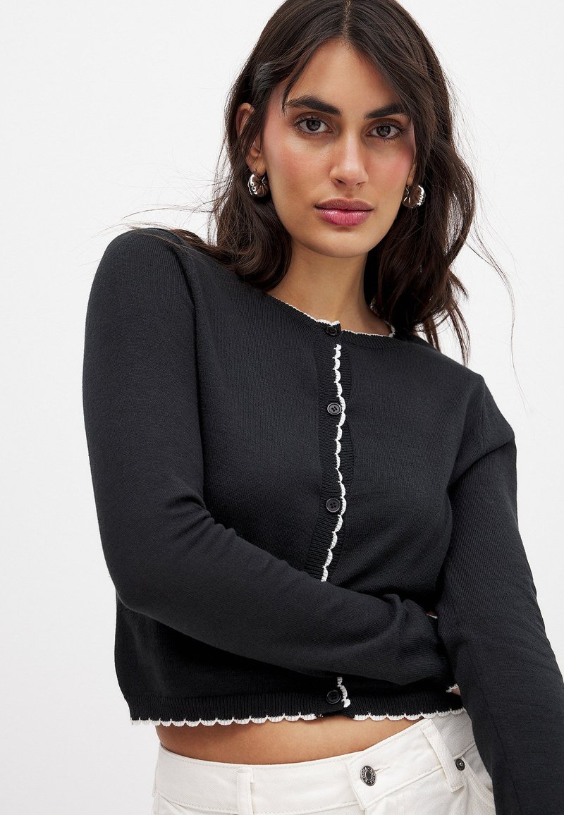 Zwarte gebreide cardigan met een ronde halslijn, witte geschulpte rand langs de randen en vijf zwarte knopen over de voorkant.