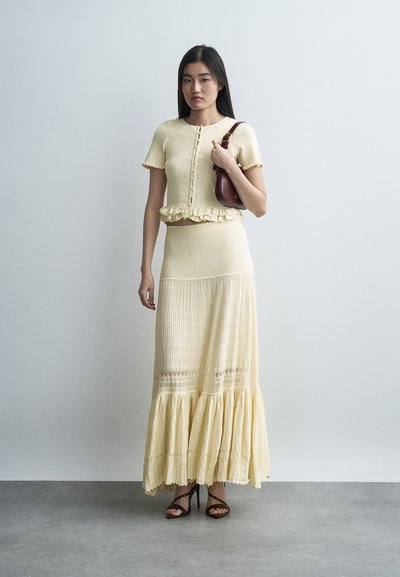 Γυναίκα στέκεται φορώντας κρεμ κοντομάνικο τοπ με κουμπιά και βολάν, μακρύ φόρεμα που ταιριάζει, μαύρα πέδιλα με λουράκια και κρατώντας μια σκούρα καφέ τσάντα ώμου.