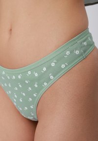 Κοντινό πλάνο πράσινου εσώρουχου με μικρό λευκό ανθικό σχέδιο, φορεμένο στην περιοχή των γοφών και της μέσης ενός ατόμου.