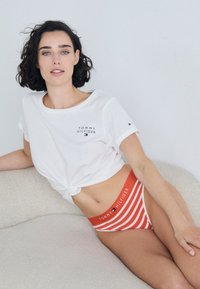 Valkoinen lyhythihainen paita, jossa on "TOMMY HILFIGER" logo. Punakuvioinen bikinit, valkoisilla ja punaisilla raidoilla, joissa on logonauha. Malli istuu vaalealla tekstuuripinnalla.