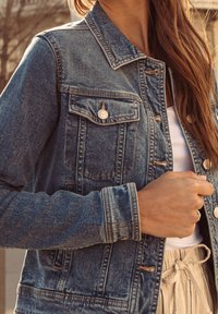 Vrouw draagt een spijkerjasje over een wit topje en beige broek, gestrikt in de taille, met lang bruin haar dat over de schouder valt.