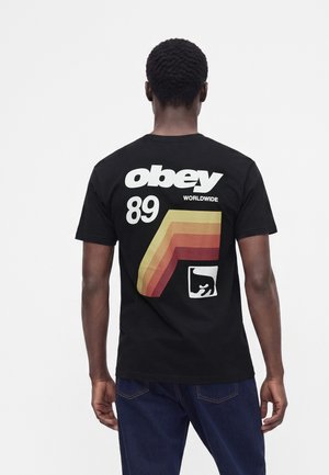 Hombre con camiseta negra con gráfico de "obey 89 worldwide" y rayas retro en la espalda, combinado con vaqueros azul oscuro.