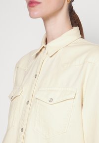 Femme portant une chemise crème claire à boutons avec deux poches poitrine et un col, cheveux attachés, et petits anneaux d'or aux oreilles.
