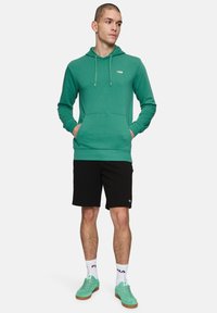 Sudadera verde con un bolsillo frontal y capucha con cordón, combinada con pantalones cortos negros. Presenta un logo blanco y zapatillas verde claro.
