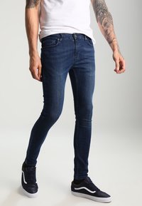 Slim jeans i mörkblå denim med en subtil struktur, utrustade med traditionella fickor och bältesloophål, tillsammans med mörka sneakers.