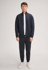 Felpa zip-up navy con polsini e vita a coste, abbinata a pantaloni jogger coordinati. T-shirt bianca sotto e sneakers bianche.