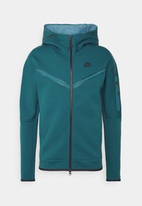 Teal zip-up Nike-huvtröja med huva, svart dragkedja, reflexiva chevron-ränder på bröstet och dragkedjeficka på vänster ärm.
