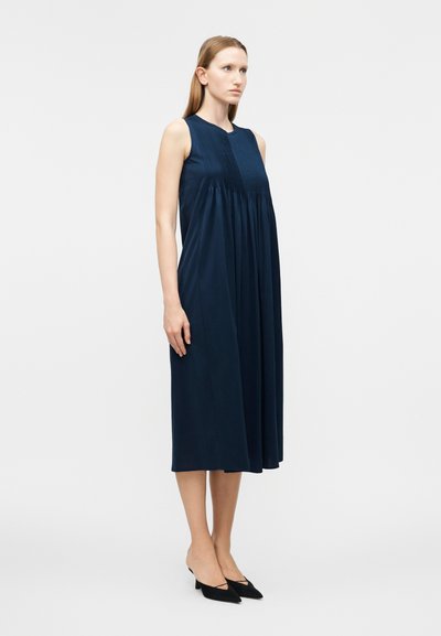 Γυναίκα που στέκεται φορώντας μαν sleeveless navy blue midi φόρεμα με πλισέ μπούστο και μαύρα μυτερά παπούτσια με χαμηλό τακούνι, σε απλό φόντο.