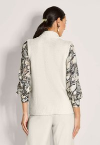 Frau mit dunklem Pferdeschwanz, die einen weißen, gestrickten ärmellosen Pullover über einer Schlangenprint-Bluse trägt, kombiniert mit hellen Hosen, von hinten gezeigt.