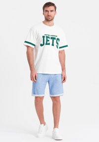 Bílý bavlněný tričko s zeleným potiskem "NEW YORK JETS" a pruhovanými rukávy, spárované se světle modrými šortkami a bílými teniskami.