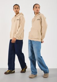 Deux personnes portant des sweats à capuche beiges avec le texte "Café", se tenant côte à côte, l'une en jeans foncés et baskets, l'autre en jeans clairs et sabots.