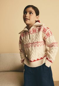 Beige stickad tröja med röda geometriska mönster, dragkedja och hög krage, kombinerad med mörka denimbyxor; mjuk textur och mysig design.