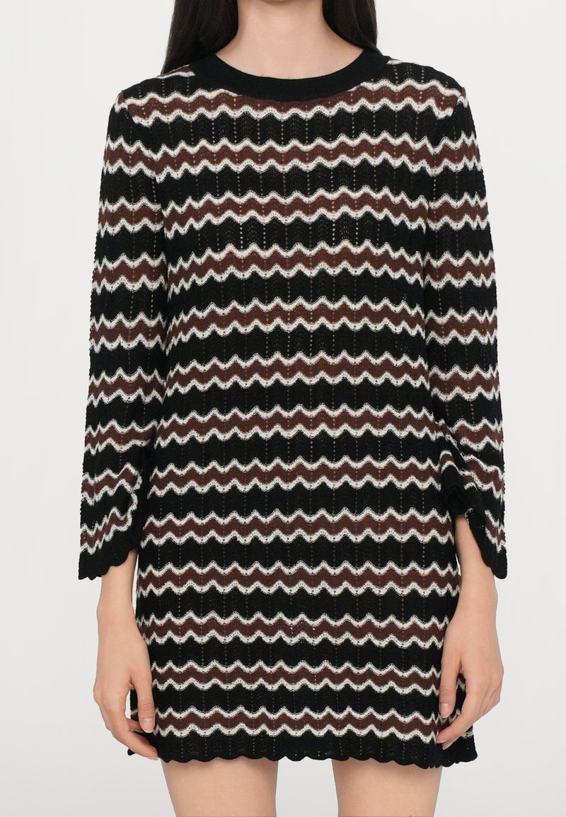 Femme portant une robe en maille à motif zigzag noir, blanc et marron avec un ourlet et des manches en feston, debout devant un fond uni.