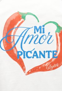 Δύο κόκκινες πιπεριές τσίλι σχηματίζουν μια καρδιά γύρω από το μπλε κείμενο "Mi Amor Picante" σε λευκό φόντο με την υπογραφή "Replay".