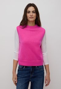 Femme aux longs cheveux bruns portant un pull sans manches rose vif sur une chemise blanche à manches longues et un jean bleu, se tenant devant un fond uni.