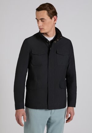 Mężczyzna w czarnej kurtce z klapami na kieszeniach i jasnoniebieskich spodniach, patrzący w dół i na bok na jednolitym tle.