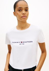 Biała bawełniana koszulka z krótkim rękawem, z nadrukiem "TOMMY HILFIGER EST. 1985" w kolorze granatowo-czerwonym na klatce piersiowej.