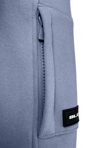 Close-up van een verticale ritszak op lichtblauwe gebreide stof met een zwarte rechthoekige patch waarop gedeeltelijk witte tekst zichtbaar is.