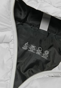 Jakkens indre med plejemærker: vandafvisende, varmesvejste sømme, genanvendt stof og genanvendt fyld, med synlig lynlås og sort foer.