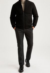 Svart quiltad jacka med hög krage, kombinerad med mörkgrå strukturerade byxor och svarta snörskor. Slät tygkvalitet med en figursydd silhuett.