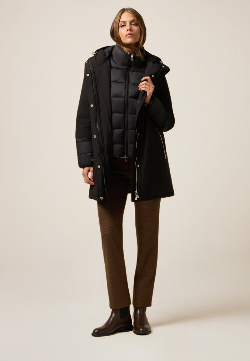 Abrigo acolchado negro con capucha, detalles de cremallera dorada, superpuesto a un chaleco puf negro. Combinado con pantalones texturizados marrones y botas oscuras.