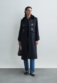 Mörkblå rock med bred krage och knäppen med togglar, kombinerad med blå jeans och svarta platta skor. Håller en liten brun handväska.