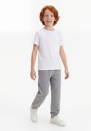 Rødhåret dreng smiler, går, bærer en ensfarvet hvid t-shirt, grå joggingbukser og hvide sneakers på en hvid baggrund.