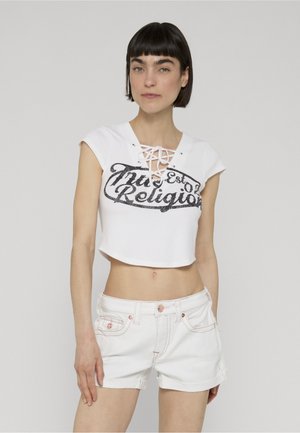 Vrouw draagt een wit cropped veter-topje met "True Religion" tekst en witte denim shorts, staand tegen een effen lichte achtergrond.