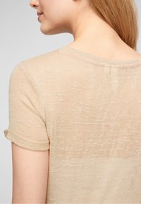 Beige stickad topp med kort ärm och texturerad yta, med rullade ärmslutar och rund halsringning. Lätt och något genomskinlig.