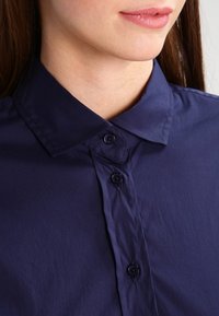 Mörkblå skjorta med knappar som har en kragdesign, mjuk textur och tre synliga svarta knappar längs knappslån.