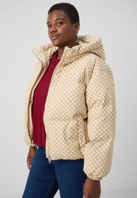 Giacca a vento trapuntata beige con motivo a diamante, dotata di cappuccio, chiusura con zip e tasche laterali. Abbinata a una maglietta bordeaux e jeans.