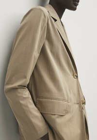 Blazer beige ajusté avec une texture lisse, fermeture à un bouton et deux poches avant. Doté d'un revers standard et d'une coupe décontractée.