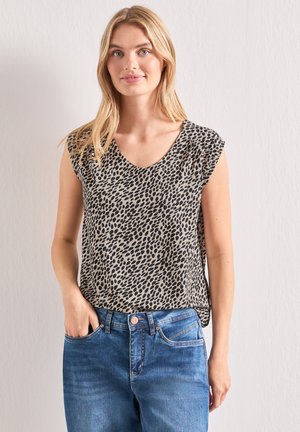 Junge Frau trägt ein ärmelloses beiges Oberteil mit schwarzen Flecken und blaue Jeans, steht vor einer schlichten hellen Wand.