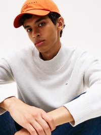 Weißer Strickpullover mit geripptem Kragen, kombiniert mit einer orangefarbenen Baseballkappe mit Logo. Das Model hat kurze dunkle Haare und sitzt.