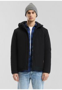 Zwarte gewatteerde jas met capuchon, gedragen over een blauw geruiten shirt, gecombineerd met lichtblauwe jeans en een grijze gebreide beanie. Eenvoudig ontwerp, geen patronen.