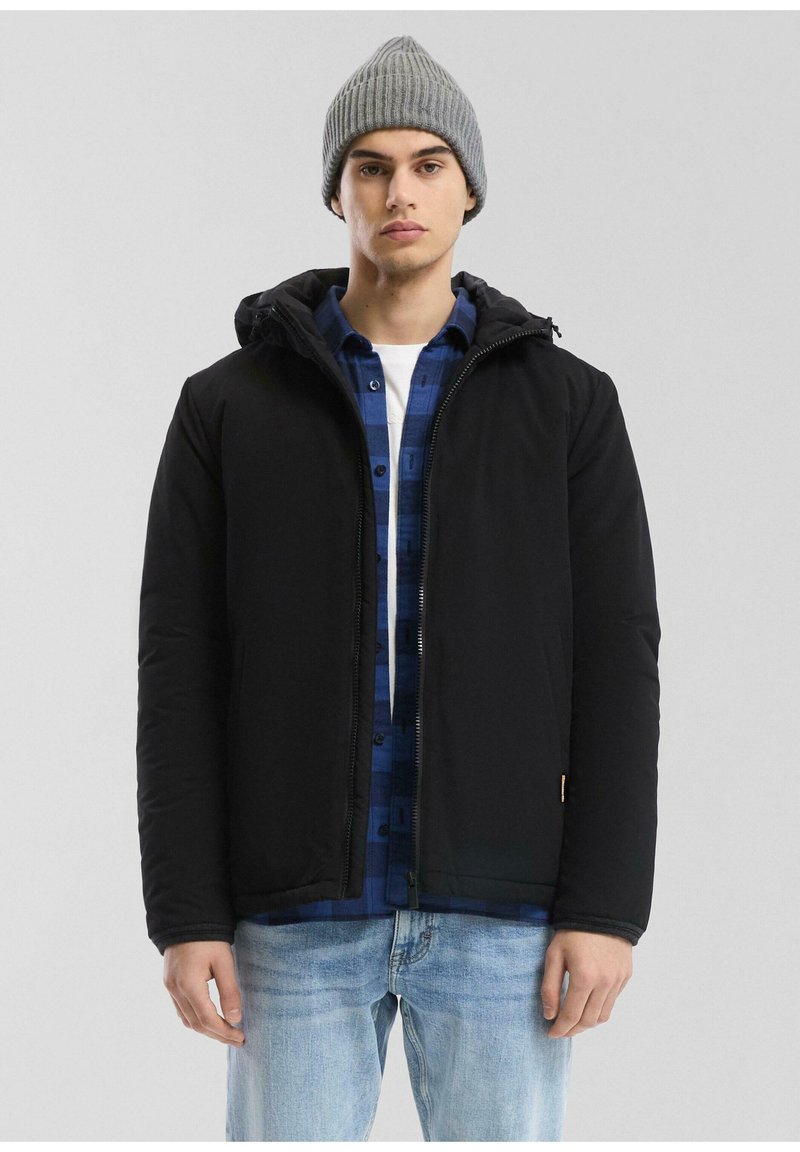 Zwarte gewatteerde jas met capuchon, gedragen over een blauw geruiten shirt, gecombineerd met lichtblauwe jeans en een grijze gebreide beanie. Eenvoudig ontwerp, geen patronen.