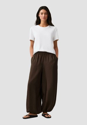 Femme aux longs cheveux bruns portant une chemise blanche à manches courtes, un pantalon ample marron foncé et des tongs noires, debout devant un fond gris.