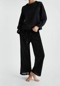 Blusa negra de manga larga con acentos de encaje floral en los hombros, combinada con pantalones negros de pierna ancha con superposición de encaje y textura semitransparente.