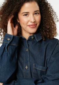 Denim shirt in dark blue with visible stitching, a buttoned collar, and a chest pocket. Features rounded cuffs with button accents.