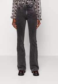 Persoon in zwarte vervaagde wijde jeans, zwart-wit gestreept shirt met lange mouwen en donkerbruine chunky schoenen, staande op een effen achtergrond.