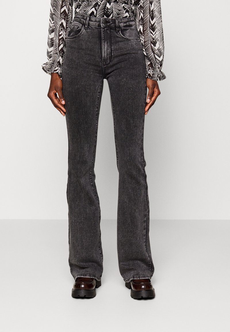 Persoon in zwarte vervaagde wijde jeans, zwart-wit gestreept shirt met lange mouwen en donkerbruine chunky schoenen, staande op een effen achtergrond.