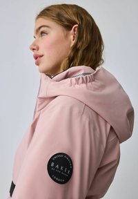 Rozā kapuce jaka ar gludu tekstūru, kurai uz piedurknes ir apaļa melna birka. Dizains ir minimālists un mūsdienīgs.