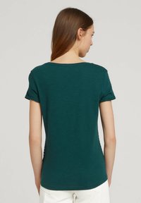 T-shirt fitted di un verde scuro, con scollatura tonda, maniche corte e una texture liscia. La parte sul retro presenta un design semplice, senza motivi o accenti visibili.