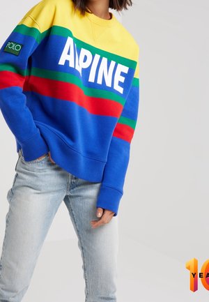 Blå sweatshirt med gule, røde og grønne striber. Har "ALPINE" i hvide bogstaver, ribbede manchetter og et logo-patch på ærmet.
