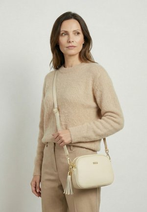 Vrouw draagt een beige trui en broek, met een crèmekleurige crossbodytas met een kwastje en gouden accenten, kijkt naar de zijkant.